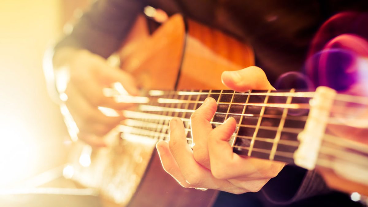 Close-up of a person playing an acoustic guitar, focusing on their left hand pressing down on the strings along the fretboard, with warm sunlight illuminating the scene.