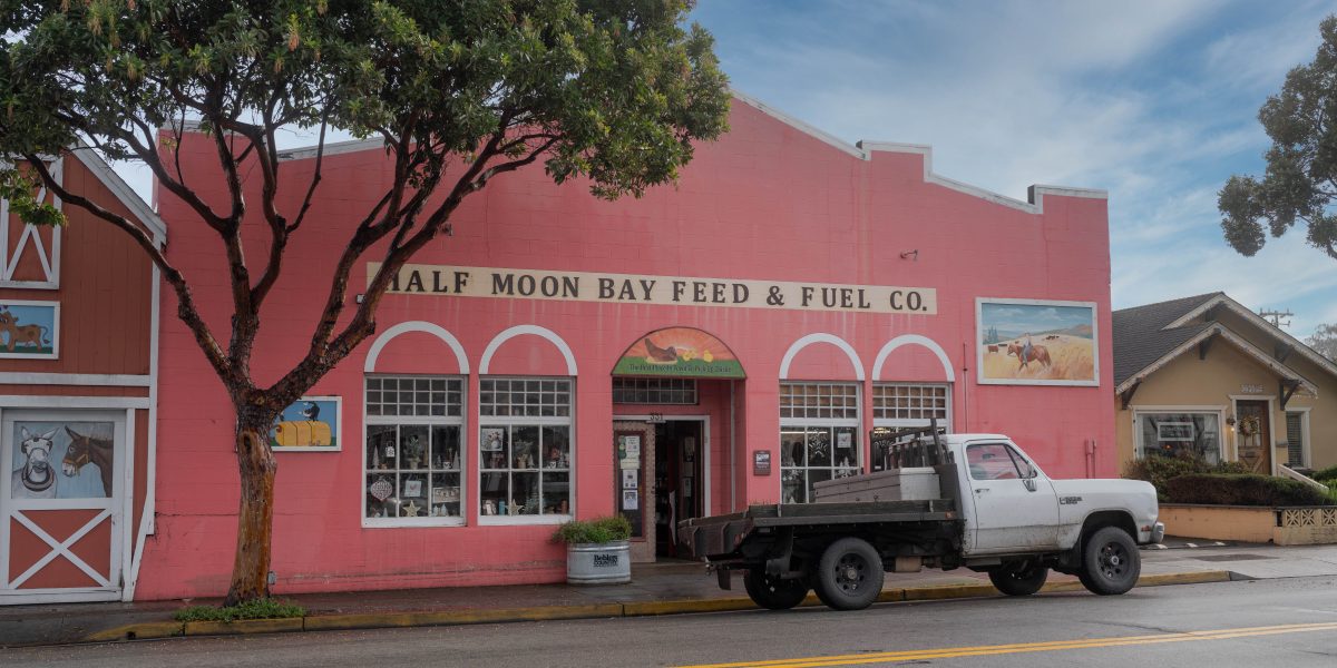 A pink building with the sign "Half Moon Bay Feed & Fuel Co." above the entrance, featuring three large windows and a central door. A white flatbed truck is parked in front of the building on a wet street. There is a large tree on the left side of the image and a smaller beige house to the right. The building has decorative paintings of horses on the left and right sides. The sky is partly cloudy.
