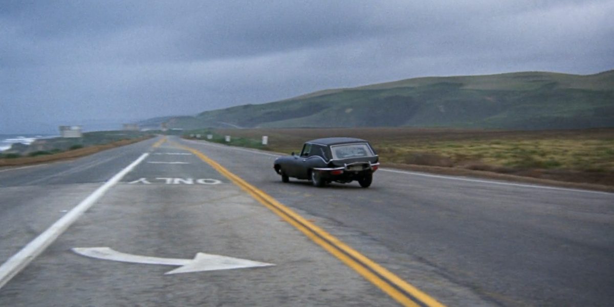 A black vintage car driving on a two-lane road with double yellow lines, heading towards a distant horizon with green hills under a cloudy sky. The road has a white arrow and the word "ONLY" painted on the left lane.