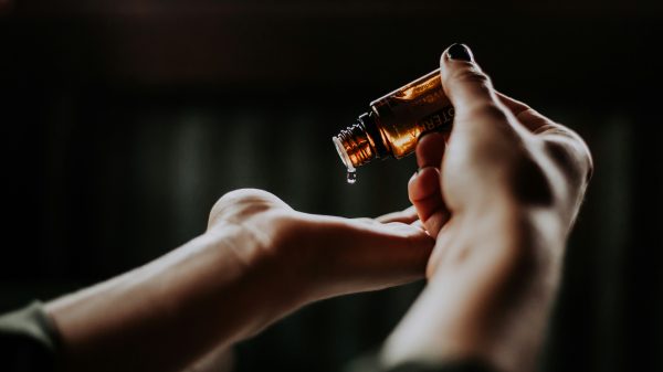 A close-up of a person holding a small amber glass bottle, pouring a drop of liquid into their open palm. The background is dark and blurred, highlighting the hands and the bottle.