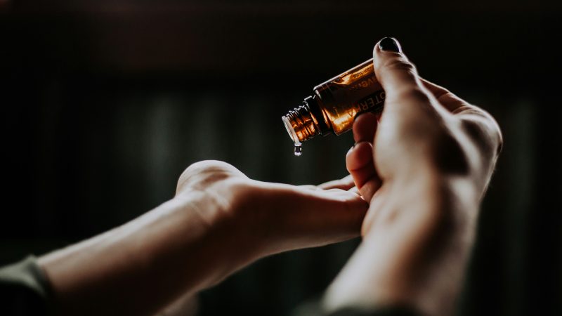 A close-up of a person holding a small amber glass bottle, pouring a drop of liquid into their open palm. The background is dark and blurred, highlighting the hands and the bottle.