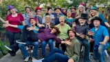 A lively group of 17 adults posing outdoors on a wooden deck, all wearing a variety of colorful and playful hats. They are smiling, laughing, and making expressive gestures, creating a joyful and energetic atmosphere. The background features green trees and a wooden fence.