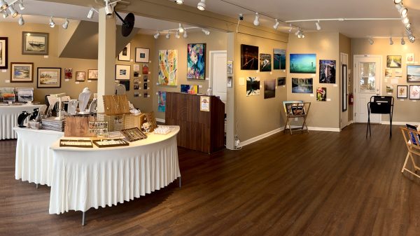 Art gallery interior with wooden flooring and beige walls adorned with various framed paintings and photographs. Two curved tables covered with white tablecloths display jewelry items such as necklaces, bracelets, and earrings on stands and trays. A wooden reception desk is positioned near the center, and several small display racks hold additional artwork. Track lighting on the ceiling illuminates the space.