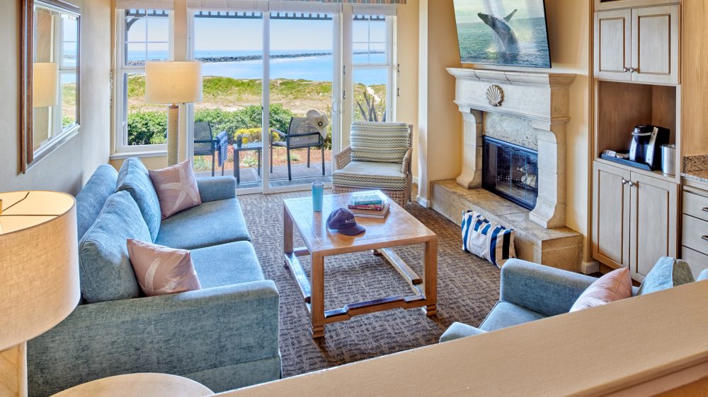 Cozy living room with blue upholstered sofa and armchair, a wooden coffee table with books, a hat, and a tumbler on it, and a patterned armchair near sliding glass doors that open to a patio with two chairs and a table. The room features a stone fireplace with a TV mounted above it displaying a whale breaching, built-in wooden cabinets with a coffee maker, and a large window offering a view of greenery and the ocean beyond. The decor includes pastel pink pillows with starfish designs and a striped valance above the windows.