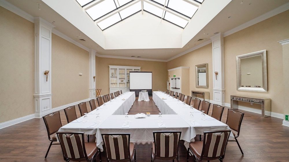 Conference room with a U-shaped arrangement of tables covered in white tablecloths, surrounded by brown chairs with striped backs. The room features beige walls, wood flooring, two large mirrors, and a skylight ceiling. A projector screen and a small table with equipment are positioned at the far end.