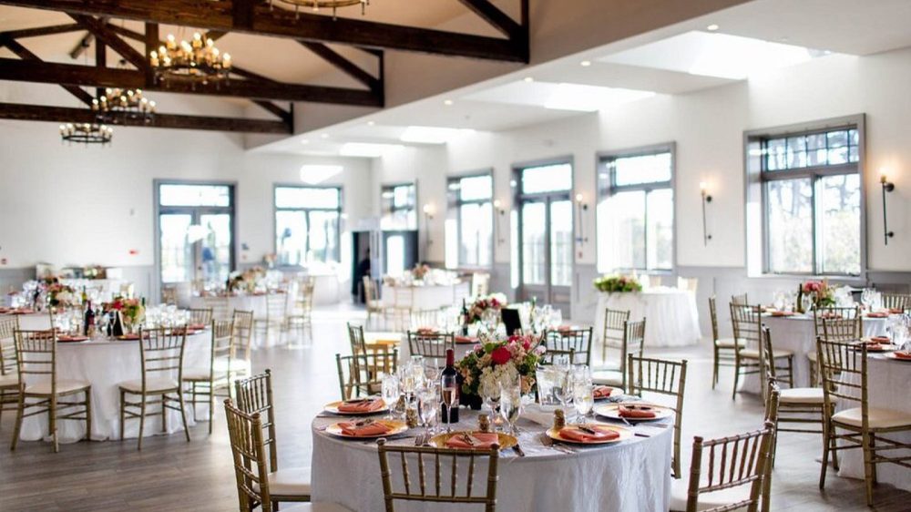 A bright, spacious event hall with wooden beams on the ceiling and multiple round tables set for a formal occasion. Each table is covered with a white tablecloth and decorated with floral centerpieces, glassware, and neatly folded orange napkins on gold chargers. Gold-colored chairs with white cushions surround the tables. Large windows and glass doors allow natural light to fill the room, enhancing the airy atmosphere. Elegant chandeliers hang from the ceiling, adding a touch of sophistication.