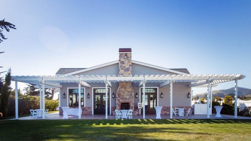 Single-story house with a stone chimney centered on the front facade, featuring a large white pergola extending across the patio. The patio is furnished with white folding chairs, small round tables, and tall cocktail tables covered with white cloth. The house has gray siding with stone accents along the lower portion of the walls and multiple glass doors and windows. The lawn in front is well-maintained, and the sky is clear and blue.