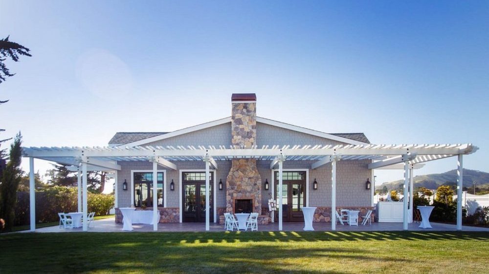 Single-story house with a stone chimney centered on the front facade, featuring a large white pergola extending across the patio. The patio is furnished with white folding chairs, small round tables, and tall cocktail tables covered with white cloth. The house has gray siding with stone accents along the lower portion of the walls and multiple glass doors and windows. The lawn in front is well-maintained, and the sky is clear and blue.