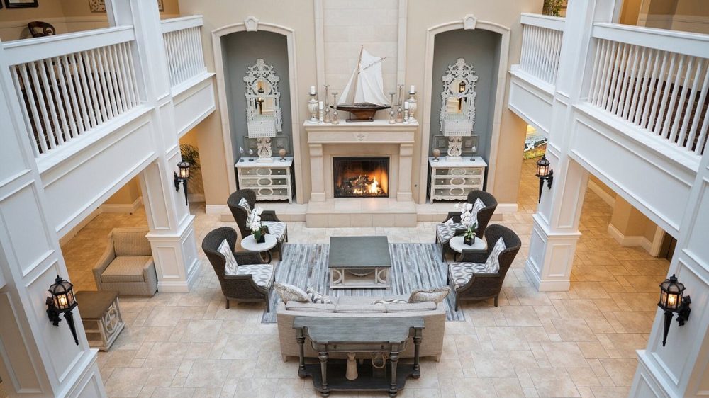 Spacious living area with a beige tiled floor and a central fireplace topped with a decorative model sailboat. The fireplace is flanked by two alcoves, each containing a vintage-style mirror and a small chest of drawers. Surrounding the fireplace are six cushioned wicker chairs with patterned pillows, arranged around a rectangular coffee table on a striped rug. A beige sofa with pillows faces the fireplace, with a wooden console table behind it. The room features white railings and columns, with wall-mounted lantern-style lights adding a warm ambiance.