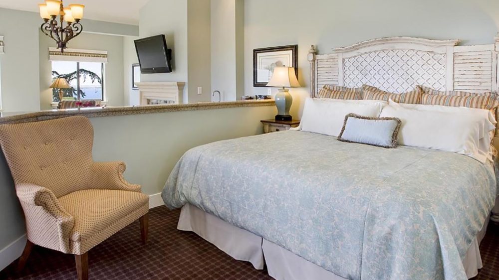 King-sized bed with a light blue patterned comforter, multiple striped and white pillows, and a white distressed wooden headboard. Beige upholstered armchair with wooden legs sits on a dark patterned carpet. A nightstand with a blue ceramic lamp and framed artwork is next to the bed. A flat-screen TV is mounted on the wall above a fireplace in the background, with a window showing a partial outdoor view. The walls are painted light blue.