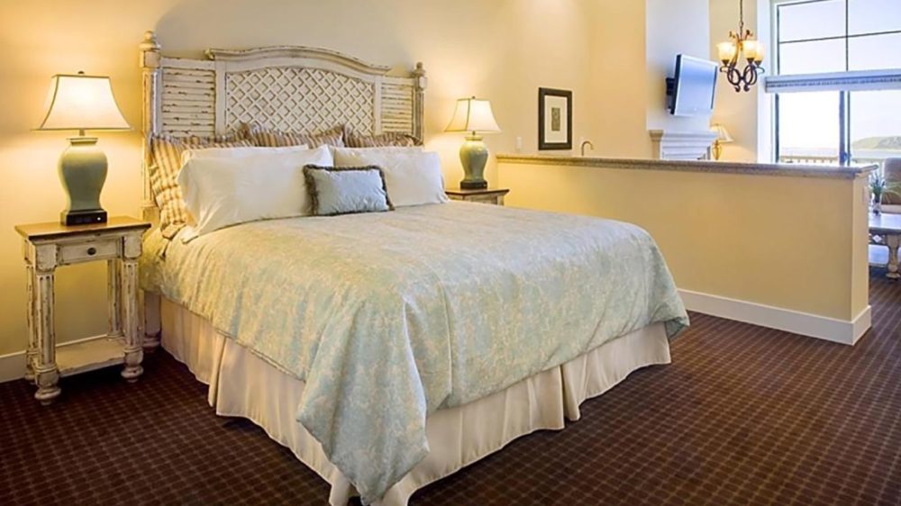 King-sized bed with a light blue patterned comforter and multiple pillows, including a small decorative pillow in front. The bed has a rustic, whitewashed wooden headboard with a lattice design. Two matching bedside tables with drawers flank the bed, each topped with a green ceramic lamp with a white lampshade. The room has beige walls and a dark brown checkered carpet. In the background, there is a half wall separating the bedroom from a sitting area with a large window, a wall-mounted TV, and a chandelier hanging from the ceiling.