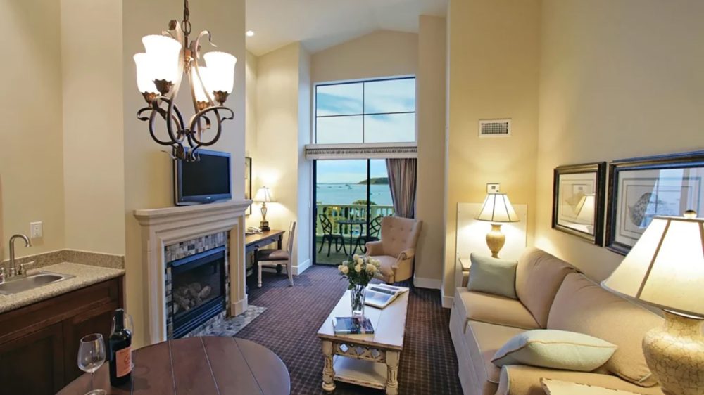 Living room with beige walls and carpet, featuring a beige sofa with pillows, a wooden coffee table with flowers and books, a fireplace with a mounted TV above it, a small desk with a chair, an armchair near a large window with a view of the ocean, and a round wooden table with a wine bottle and glass. Several lamps and a chandelier provide lighting.