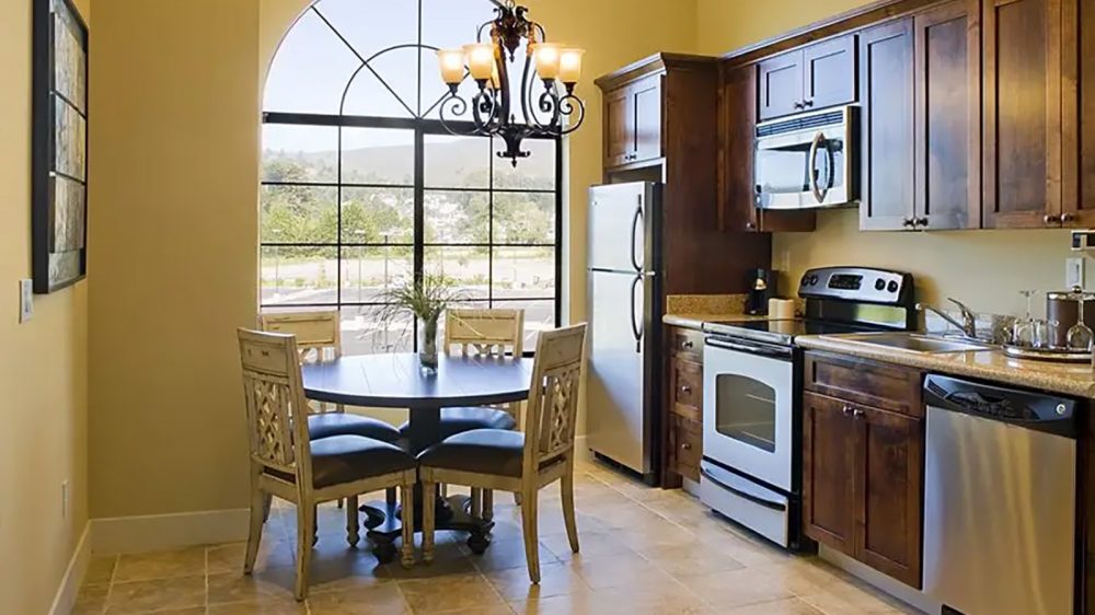 Kitchen with dark wood cabinets, stainless steel appliances including a refrigerator, oven, microwave, and dishwasher. A round dining table with four wooden chairs is positioned near a large arched window, with a decorative chandelier hanging above the table. The floor is tiled, and the walls are painted a warm beige color.