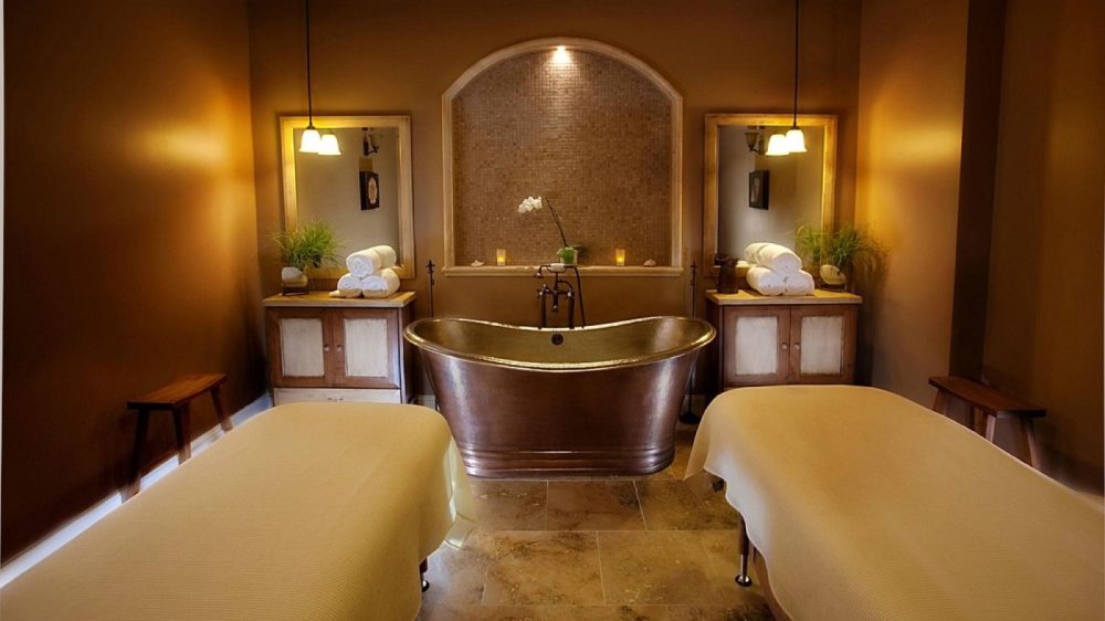 A warm-toned spa room featuring two massage tables covered with beige sheets, a central copper bathtub, and two wooden cabinets with mirrors above them. Each cabinet holds neatly rolled white towels and a small potted plant. Soft pendant lights hang above the cabinets, and the back wall has a recessed arch with mosaic tiles, a single spotlight, and small candles. The floor is tiled in natural stone.