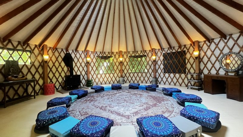 A spacious yurt interior with a circular arrangement of blue and gray meditation cushions on a large round patterned rug. The yurt has wooden lattice walls and a high ceiling with wooden beams radiating from a central skylight. There are wall-mounted lights evenly spaced around the perimeter, a black wood stove on the left, a wooden desk with a round mirror on the right, and a small table with decorative items on the far left. The atmosphere is calm and inviting, suitable for group meditation or gatherings.