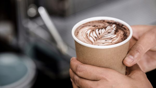 A close-up of hands holding a brown paper cup filled with hot chocolate topped with latte art in the shape of a leaf. The background is blurred, highlighting the cup and the hands.