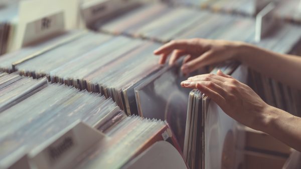 Hands browsing through a collection of vinyl records organized in rows with labeled dividers. The scene has a soft, warm lighting and a slightly blurred background.