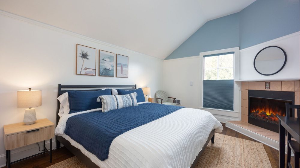 Bedroom with a large bed featuring white and navy blue bedding, two bedside tables with lamps, a woven rug, a fireplace with a round mirror above it, a window with a blue shade, and three framed beach-themed pictures on the wall. The walls are white with a blue accent on the upper part of the slanted ceiling.