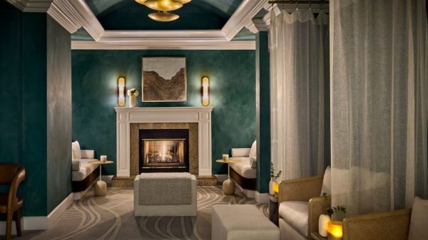 A cozy, elegant room with deep green walls and a white fireplace at the center, featuring a lit fire. Above the fireplace hangs a framed abstract artwork. Two wall sconces with warm lighting flank the artwork. The ceiling is vaulted and painted in the same green tone, with white crown molding and a gold chandelier. On either side of the fireplace are white cushioned benches with small round side tables holding candles. In the foreground, there are beige cushioned ottomans and armchairs separated by sheer white curtains, with small wooden tables holding glowing candles and small plants. The carpet has a subtle, flowing pattern in neutral tones.