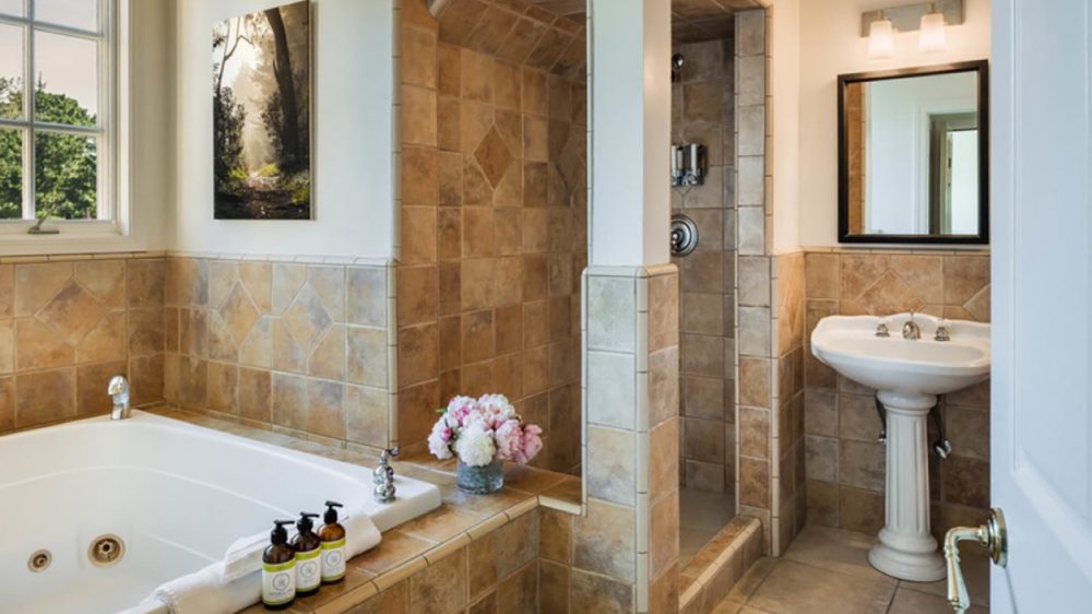 Bathroom with beige and brown tiled walls and floor, featuring a white jetted bathtub with bath products and a towel on its edge, a small vase of flowers on the tiled ledge, a walk-in tiled shower, and a white pedestal sink with a mirror and light fixture above it. A window and a nature-themed wall art are visible on the left side.