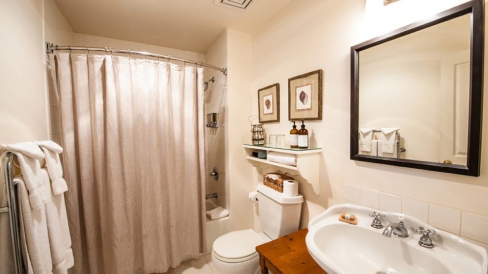 Small bathroom with a white toilet, a white pedestal sink, and a wooden stool beside the sink. A beige shower curtain hangs on a curved rod over the bathtub. White towels are neatly folded on a metal towel rack. Above the toilet, a white shelf holds toiletries and two framed pictures with seashell designs. A large rectangular mirror with a dark frame is mounted above the sink, illuminated by a wall light. The walls and floor tiles are light-colored, creating a clean and cozy atmosphere.