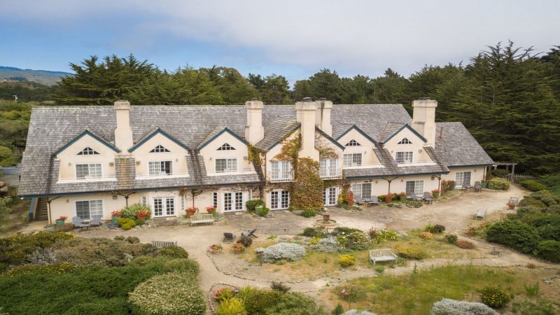 Large two-story house with a gray shingled roof and cream-colored walls, featuring multiple chimneys and dormer windows. Ivy climbs the central section of the house. The front yard includes a circular garden with a small fountain, various plants, flower pots, benches, and outdoor seating areas. The house is surrounded by dense green trees and shrubs.