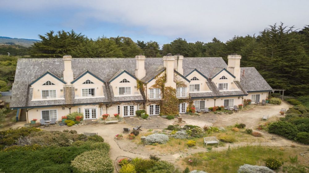 Large two-story house with a gray shingled roof and cream-colored walls, featuring multiple chimneys and dormer windows. Ivy climbs the central section of the house. The front yard includes a circular garden with a small fountain, various plants, flower pots, benches, and outdoor seating areas. The house is surrounded by dense green trees and shrubs.