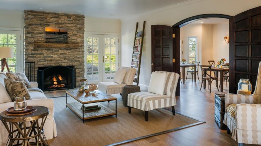 Living room with a stone fireplace, a white sofa with patterned pillows, two striped armchairs, a wooden coffee table, and a side table with a candle. The room features wooden floors, a large area rug, and dark wooden double doors leading to a dining area with wooden chairs and tables. A ladder-style magazine rack leans against the wall near French doors that open to an outdoor space.
