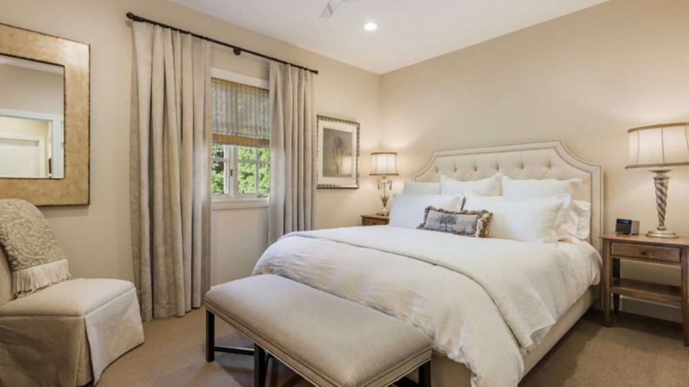 Bedroom with a large upholstered bed featuring a tufted headboard, white bedding, and multiple pillows. A beige bench with dark wooden legs is placed at the foot of the bed. Two wooden nightstands with matching lamps flank the bed. Beige curtains and a woven window shade cover the window. A beige upholstered chair with a patterned throw is positioned near a large framed mirror on the wall. The room has neutral beige walls and carpet, with a ceiling fan and recessed lighting.