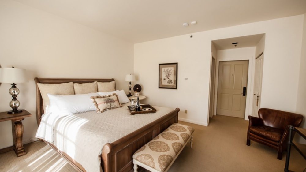 Bedroom with a wooden bed frame and beige bedding, featuring multiple pillows and a decorative cushion. A wooden bench with patterned upholstery is placed at the foot of the bed. There are two bedside tables with lamps, a framed artwork on the wall, a brown leather armchair, and a beige carpeted floor. The room has a neutral color palette with cream walls and a beige door in the background.
