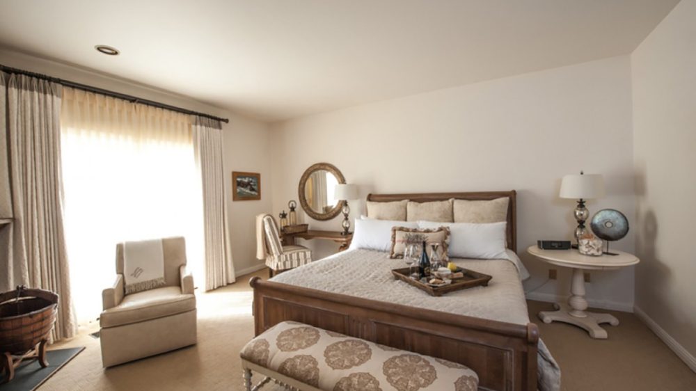 A cozy bedroom with a wooden bed frame and a beige quilted bedspread. The bed has multiple pillows and a decorative tray with wine glasses and a bottle. At the foot of the bed is an upholstered bench with a floral pattern. To the right, there is a round white side table with a lamp, a decorative globe, and a clock. On the left side, a beige armchair is positioned near a large window with sheer and heavy curtains. A small wooden desk with a striped chair and a round mirror above it is also visible in the corner. The room has neutral tones and soft lighting.