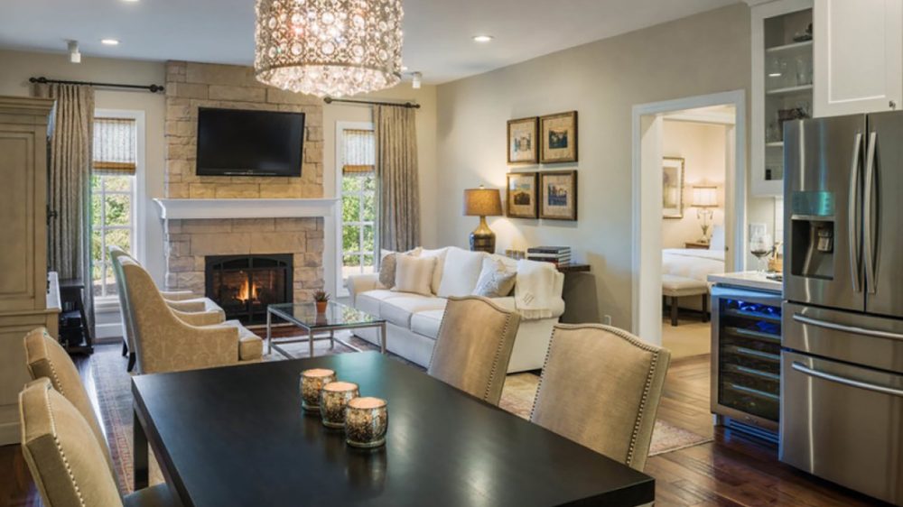 Open-concept living and dining area with a dark wood dining table and beige upholstered chairs, a white sofa with cushions, a stone fireplace with a mounted TV above, two armchairs, a glass coffee table, a decorative chandelier, and a stainless steel refrigerator visible near the kitchen entrance.