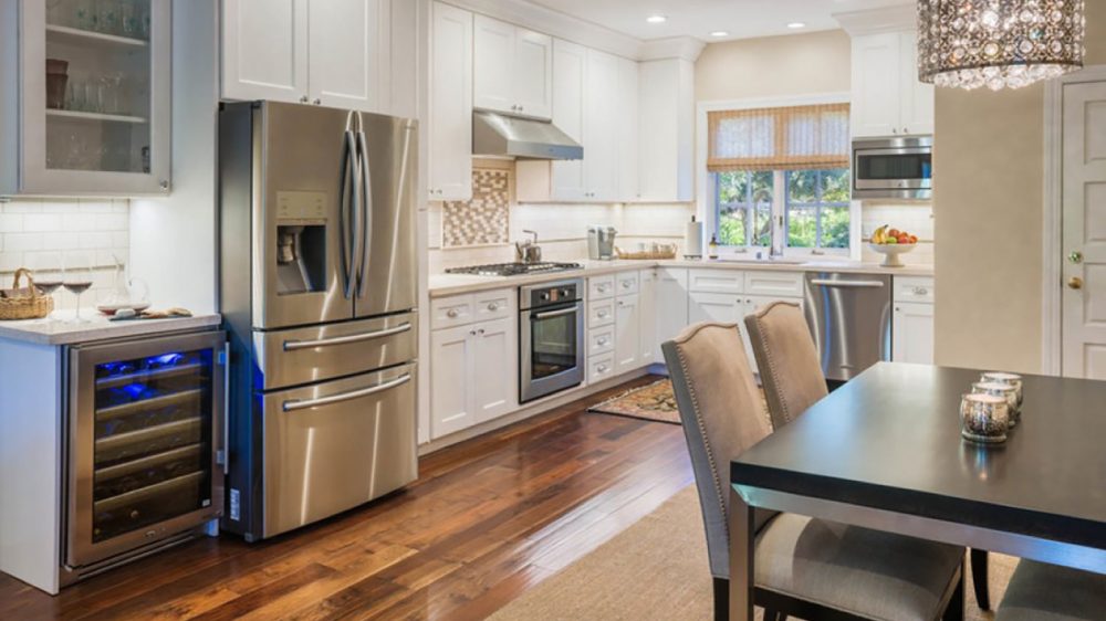 Modern kitchen with white cabinetry, stainless steel appliances including a refrigerator, oven, microwave, dishwasher, and a wine cooler. The space features hardwood floors, a black dining table with beige upholstered chairs, a crystal chandelier, and a window with a bamboo shade. The countertops are light-colored, and there is a small fruit bowl on the counter near the window.