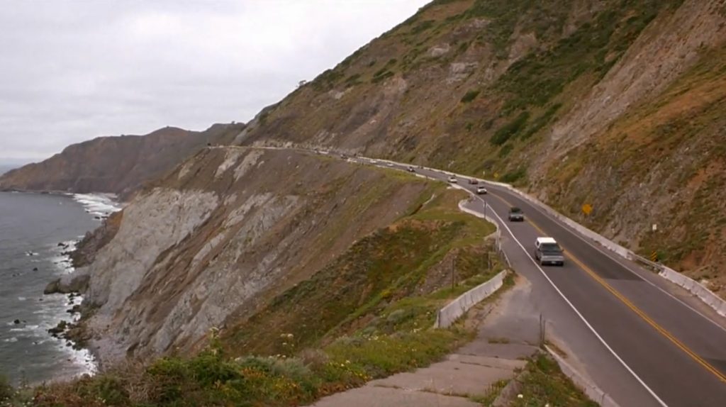 A coastal highway winding along steep cliffs with several cars driving in both directions. The cliffs are rocky with patches of greenery, and the ocean waves crash against the base of the cliffs. The sky is overcast.
