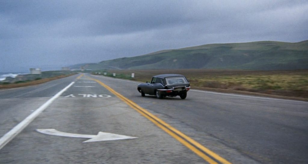 A black vintage car driving on a two-lane road with double yellow lines, heading towards a distant horizon with green hills under a cloudy sky. The road has a white arrow and the word "ONLY" painted on the left lane.