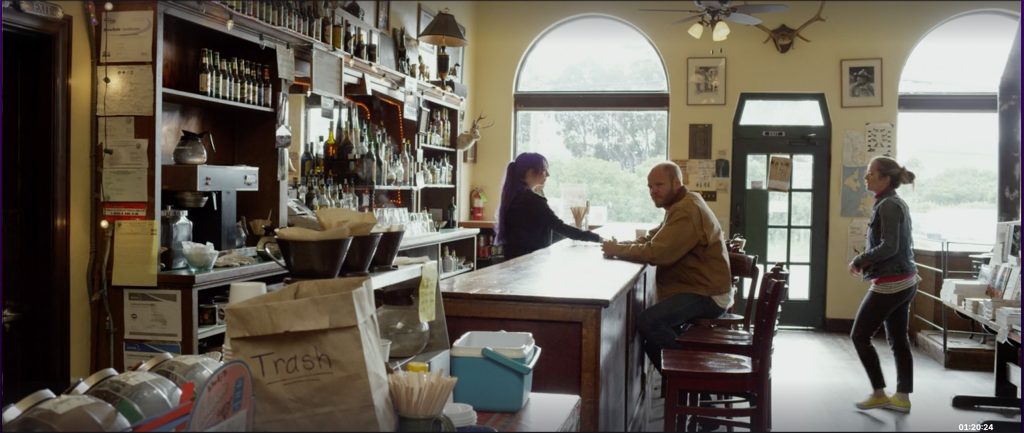 A rustic bar interior with wooden shelves stocked with various bottles and glassware. A man in a brown jacket sits on a wooden bar stool at the counter, facing a woman with long purple hair standing behind the bar. Another woman in casual clothing walks past the bar on the right side. The room features a large arched window letting in natural light, a ceiling fan with lights, and mounted animal heads on the wall. A paper bag labeled "Trash" and a container with straws are visible in the foreground.