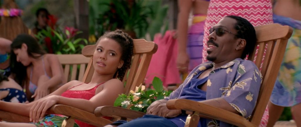 A woman in a red top and colorful skirt and a man wearing sunglasses, a blue shirt, and a patterned short-sleeve button-up shirt are sitting and relaxing on wooden lounge chairs outdoors, surrounded by greenery and flowers. The woman looks to the side while the man appears to be talking or laughing.