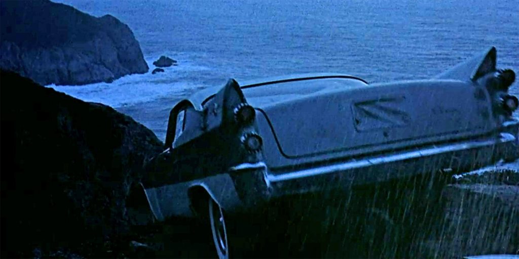A dark-colored vintage car is precariously hanging off the edge of a rocky cliff overlooking the ocean during a rainy, dimly lit scene. The car's rear end is tilted downward, and the coastline with waves crashing against rocks is visible in the background.