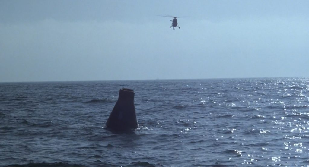 A partially submerged space capsule floats in the ocean with a helicopter hovering above it against a clear sky. The water is slightly choppy with sunlight reflecting off the surface.