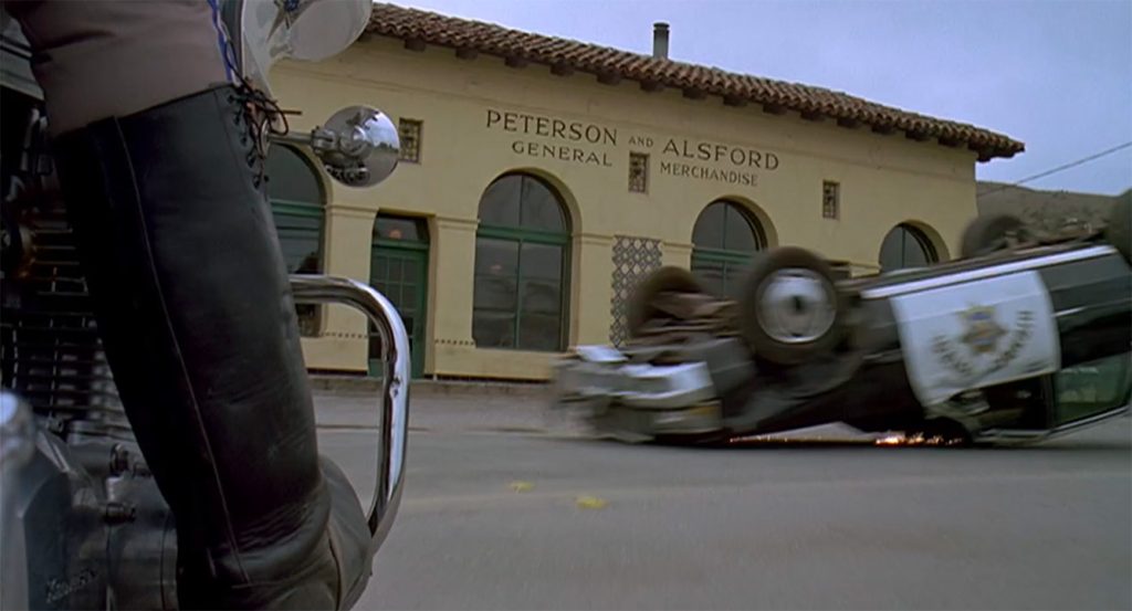 A police car is flipped upside down on a street in front of a building with arched windows and green doors. The building has a sign that reads "Peterson and Alsford General Merchandise." In the foreground, a person wearing black leather boots is sitting on a motorcycle.